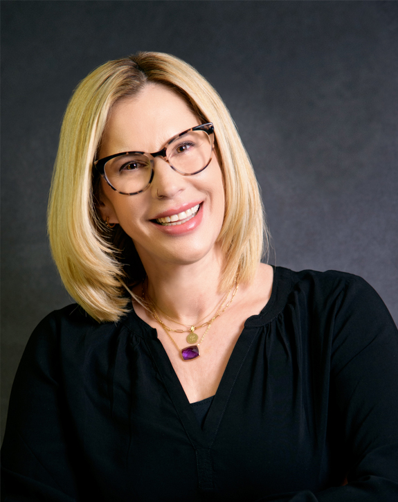 A woman with blonde hair wearing glasses and smiling.